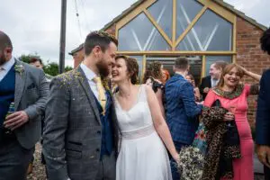 A bride and groom, adorned with confetti, share a joyful moment outside a brick building. They are surrounded by smiling, well-dressed guests.