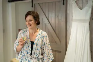 A woman in a floral robe smiles while holding a drink next to a hanging white dress.