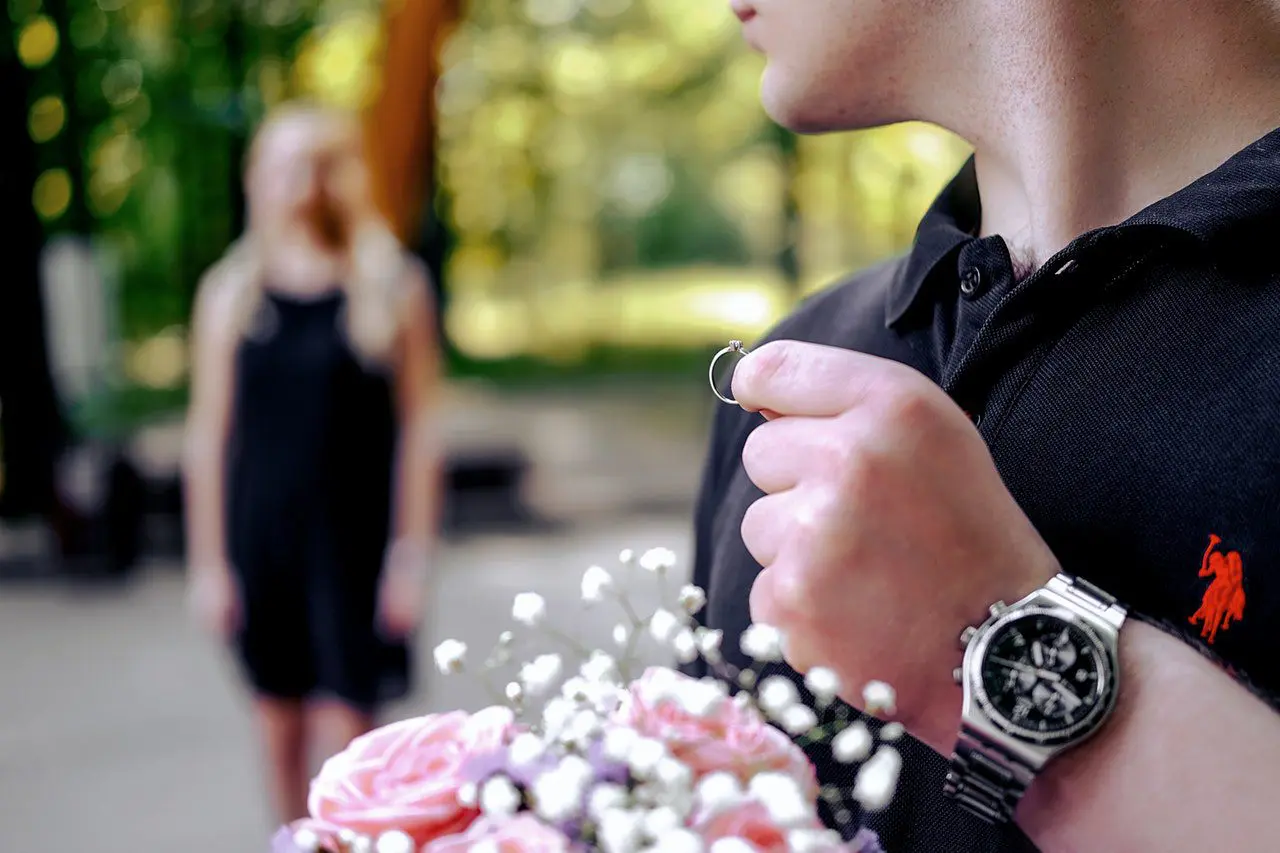 A person in a black polo shirt holds a ring and a bouquet of flowers, while another person in a black dress stands in the blurred background in a park setting.