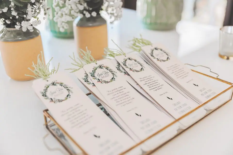 A tray holds several elegant wedding programs decorated with floral designs, placed next to potted plants on a white table.