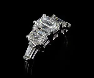 A silver ring with a rectangular center diamond flanked by four smaller rectangular diamonds on a black background.