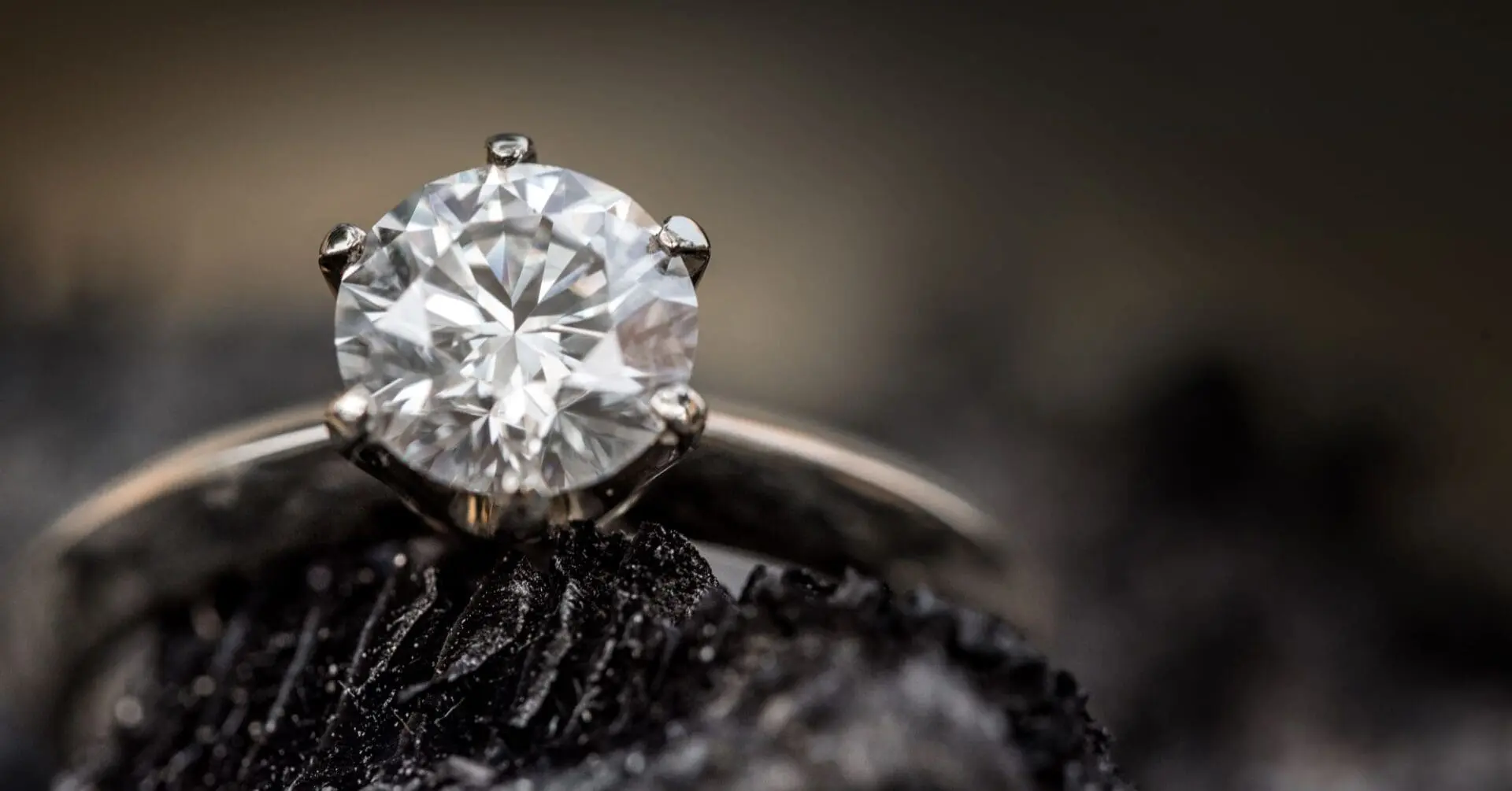 Close-up of a silver ring featuring a large, round-cut diamond set in prongs, placed on a textured, dark surface.