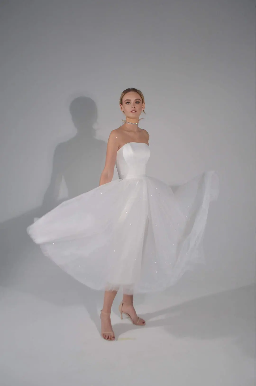 A woman in a strapless, white tulle dress stands in a studio setting. She has short hair and is wearing beige high heels.