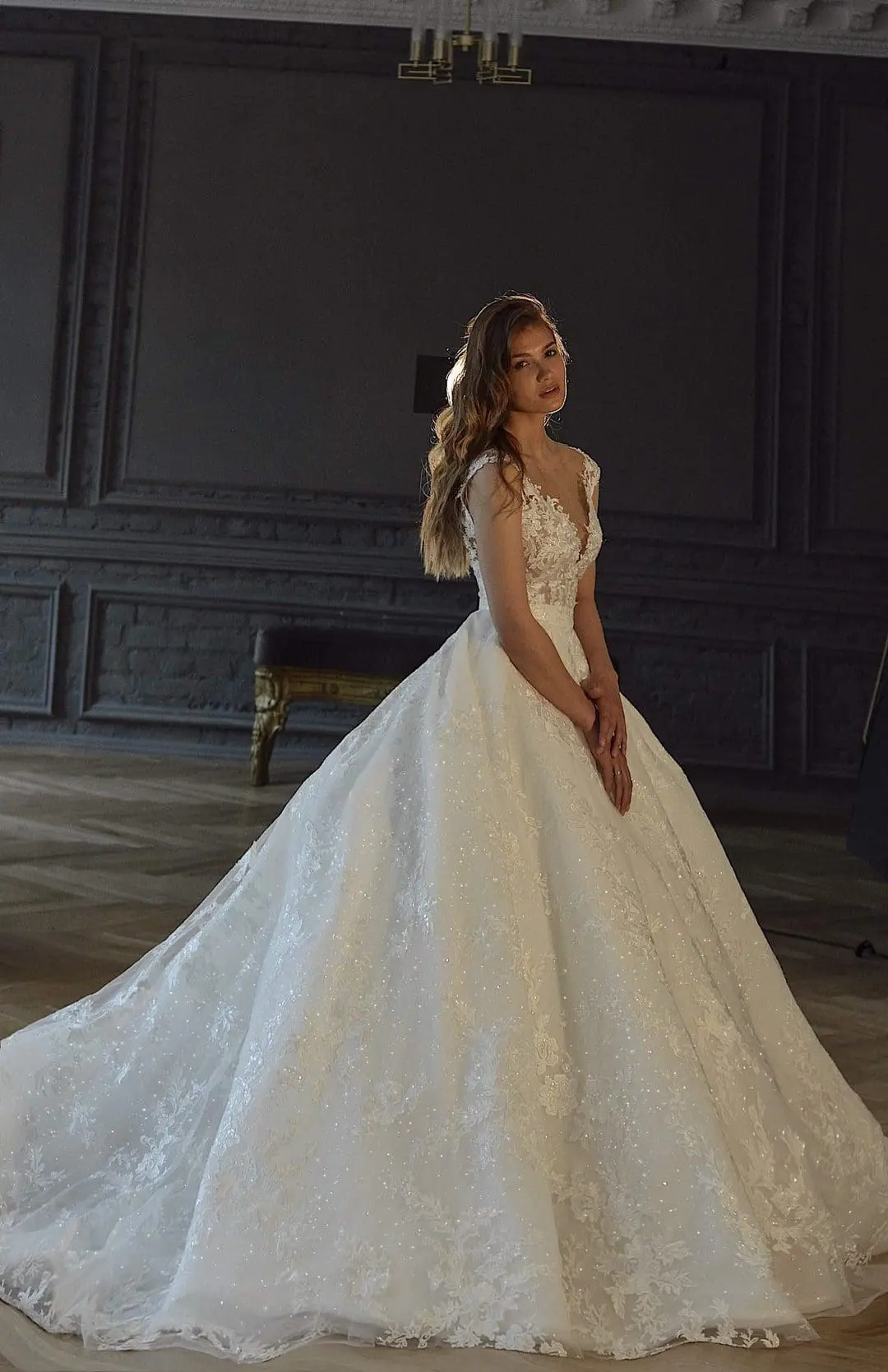 A woman stands in an elegant ballroom wearing a white bridal gown with lace detailing. She is posing with her hands clasped, looking to the side. The room features ornate wood paneling and a wooden floor.