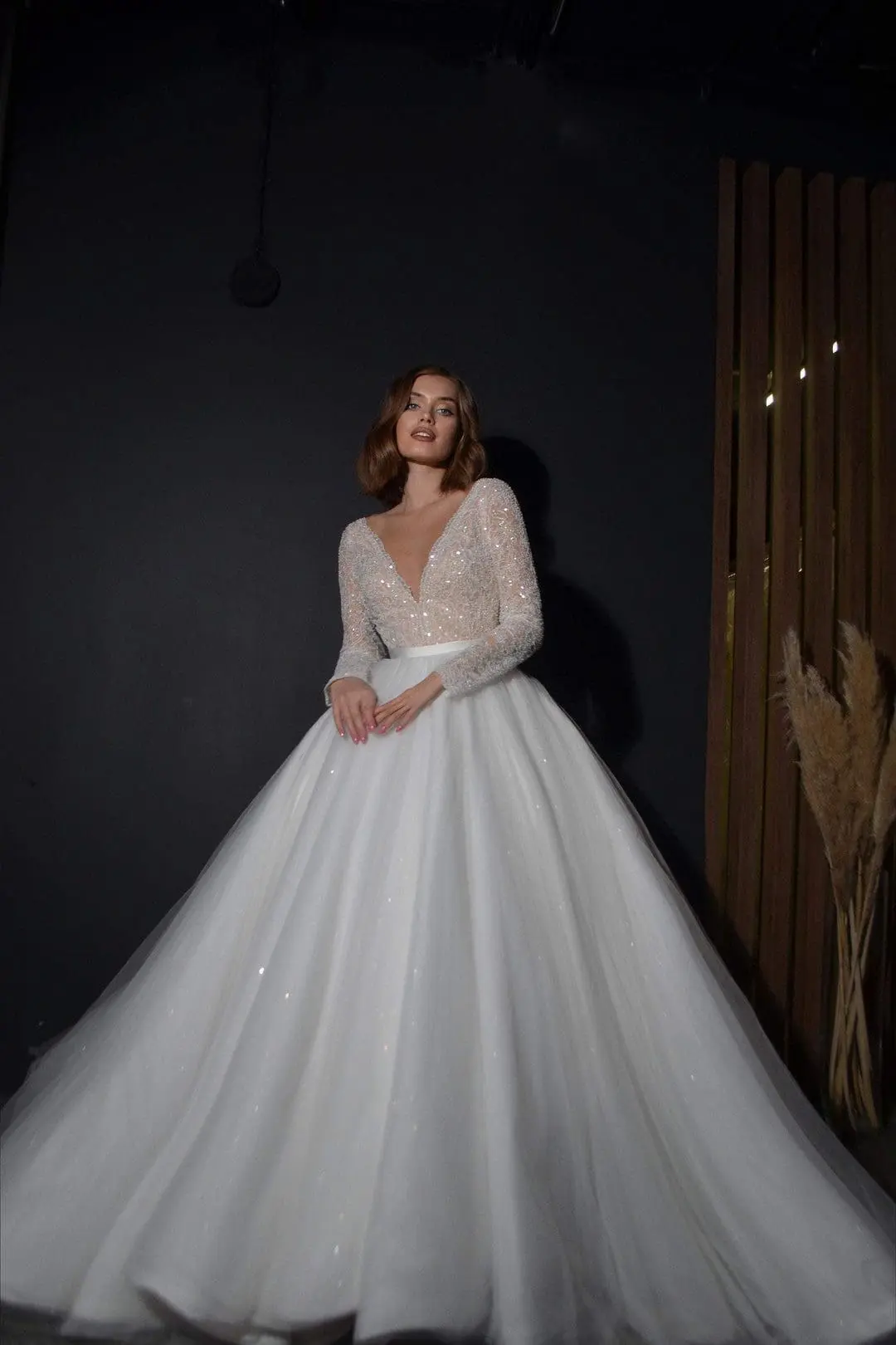 A woman in a sparkling, long-sleeved white wedding dress poses against a dark backdrop.