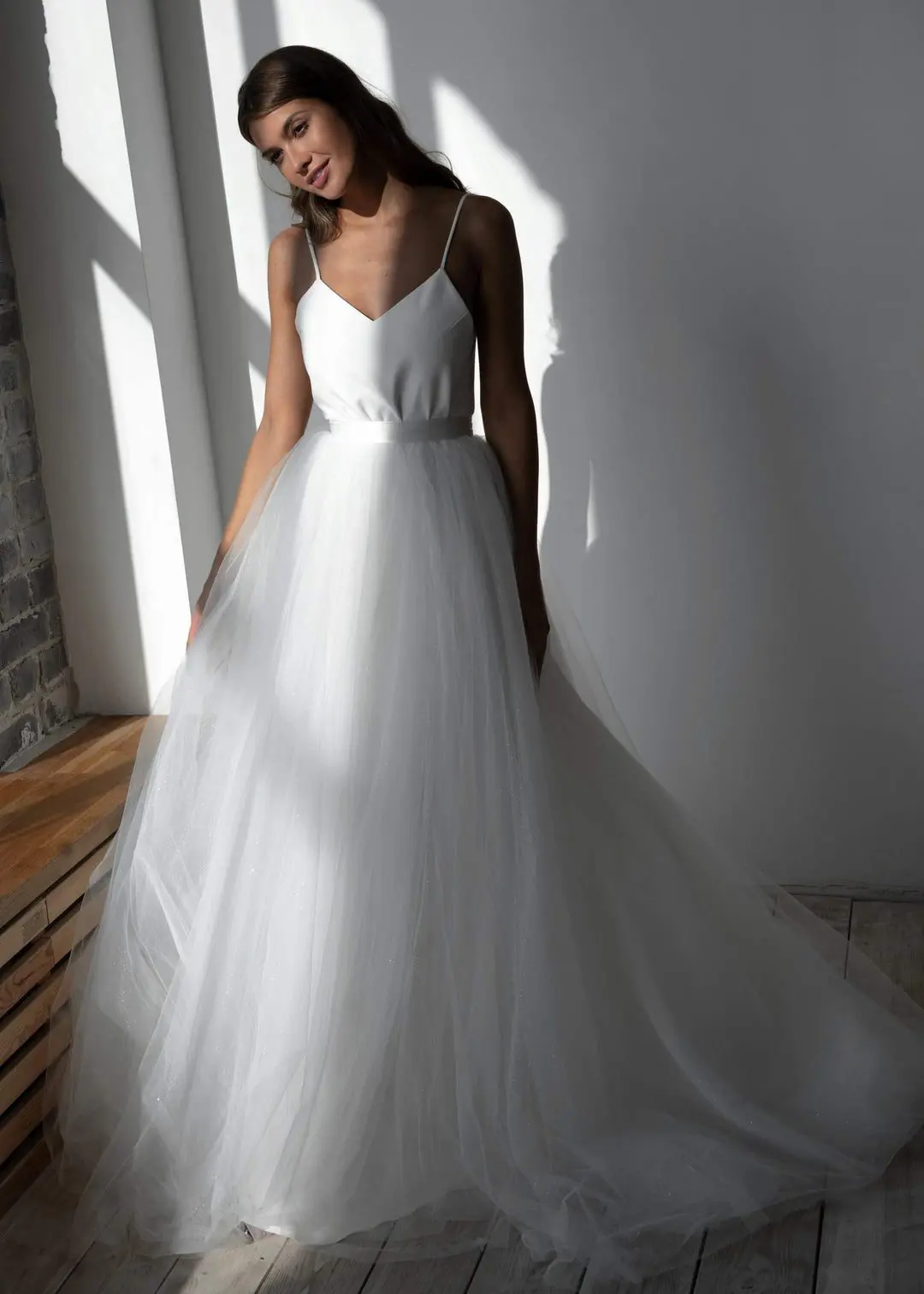 A woman in a white wedding dress with a tulle skirt and spaghetti straps stands near a sunlit window.