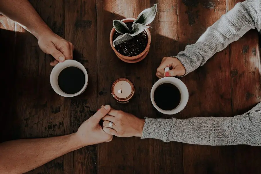 Two people holding hands and each holding a cup of coffee at a wooden table with a small potted plant and a lit candle.