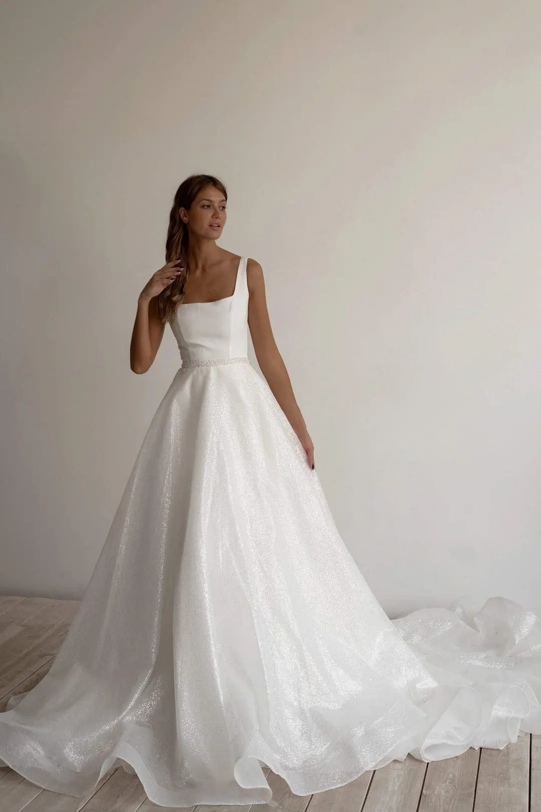 A woman in a white sleeveless wedding gown stands on a light-colored wooden floor with a long train spreading out behind her.
