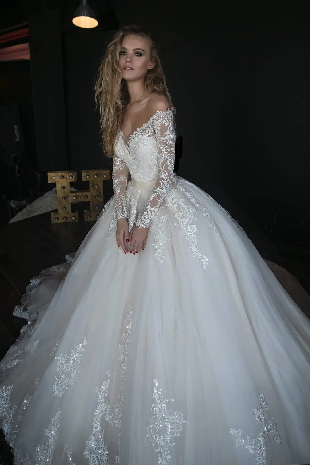 A woman in a white, lace, off-the-shoulder ball gown wedding dress stands indoors against a dark background. She has long, wavy hair and is gazing slightly downward.