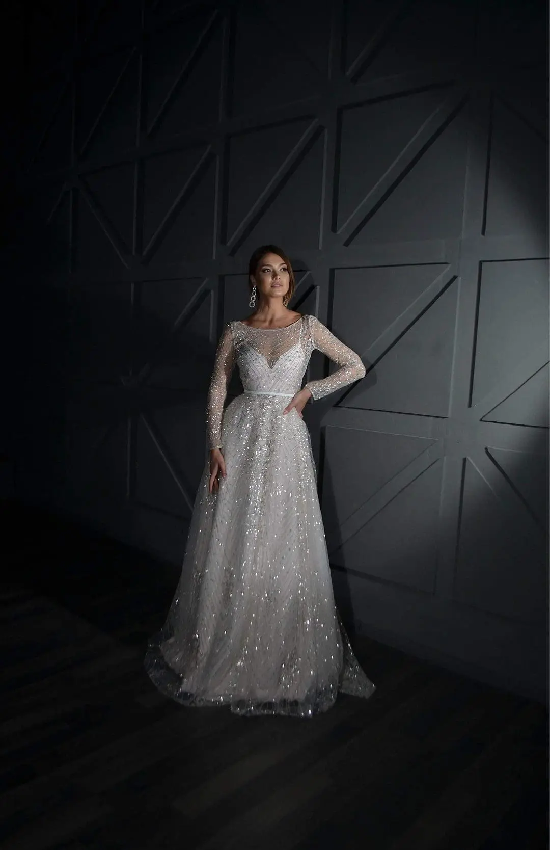 A woman in a sparkling, long-sleeved gown stands against a geometric patterned wall, her left hand resting on her hip. The scene is dimly lit.