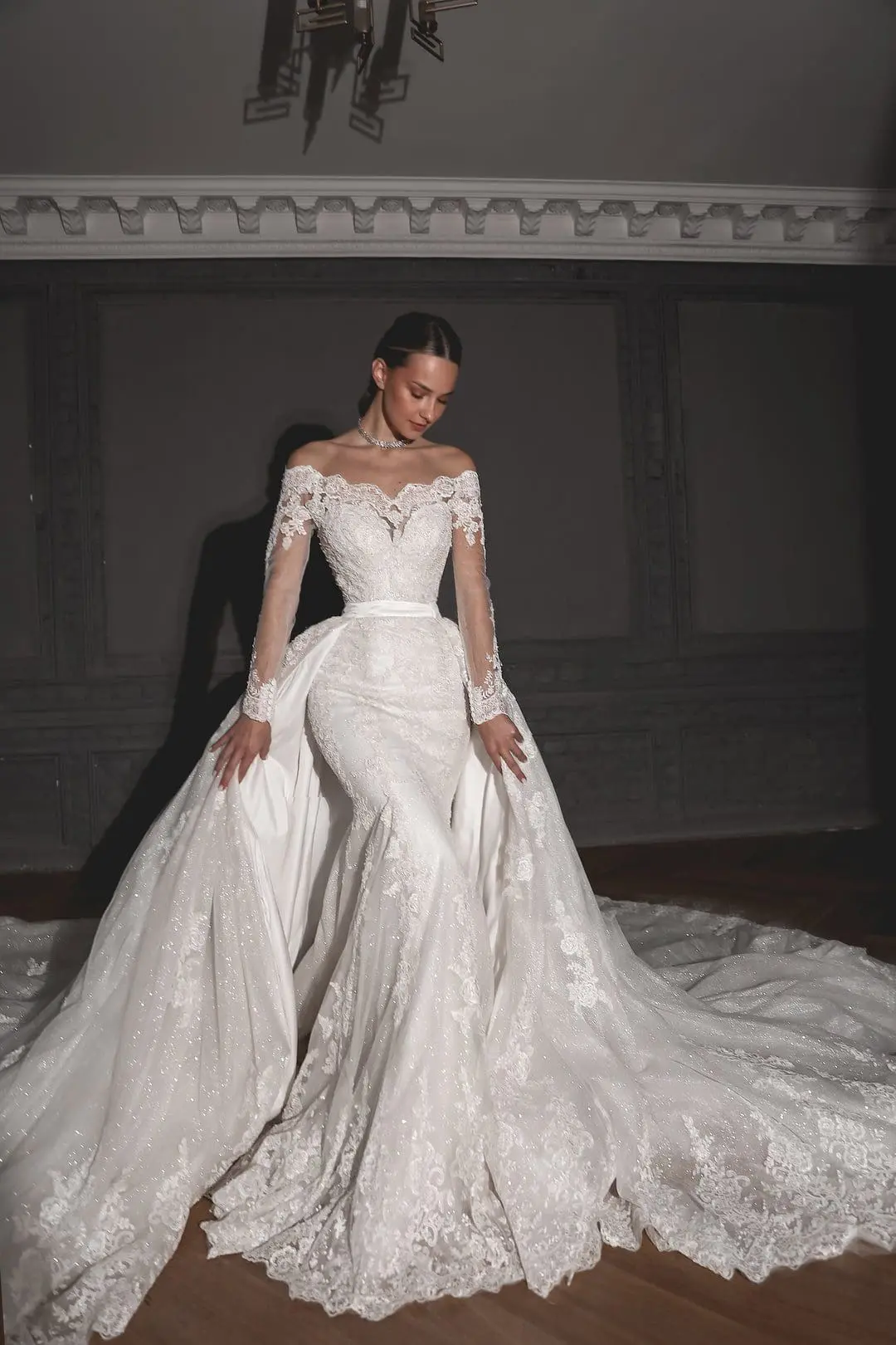 A person is wearing an elaborate off-shoulder wedding dress with lace details, long sleeves, and a voluminous train, posing indoors against a dark background.