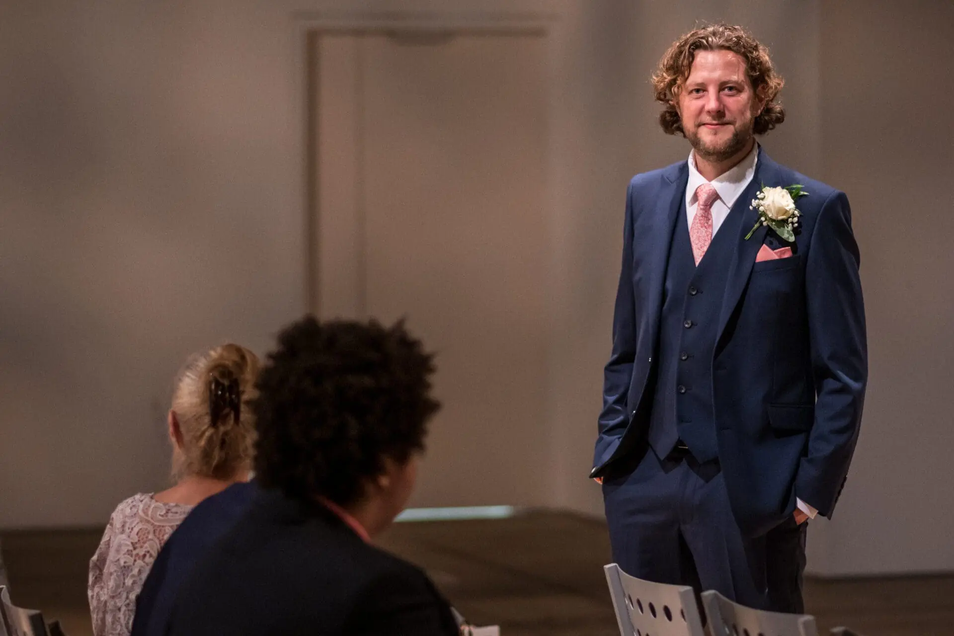 A man in a blue suit with a boutonniere stands in a room with white walls. He faces the camera, while people seated in the foreground observe him.