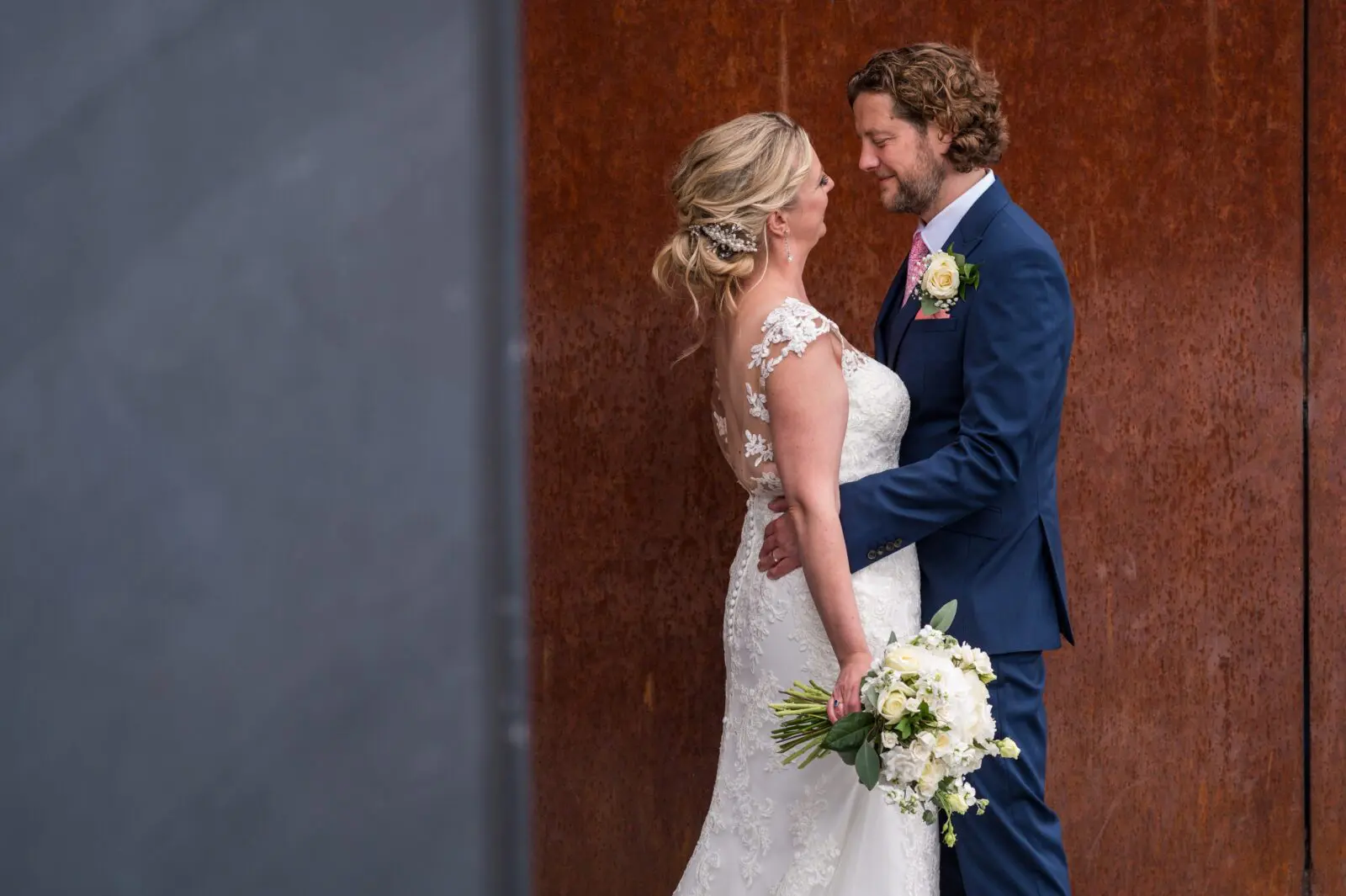 A bride in a lace wedding dress and groom in a blue suit stand facing each other, smiling, with the bride holding a bouquet of white flowers. A dark and rust-colored background is behind them.