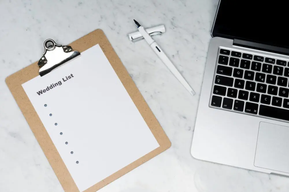 A clipboard with a blank wedding list, pen, and an open laptop on a marble surface.