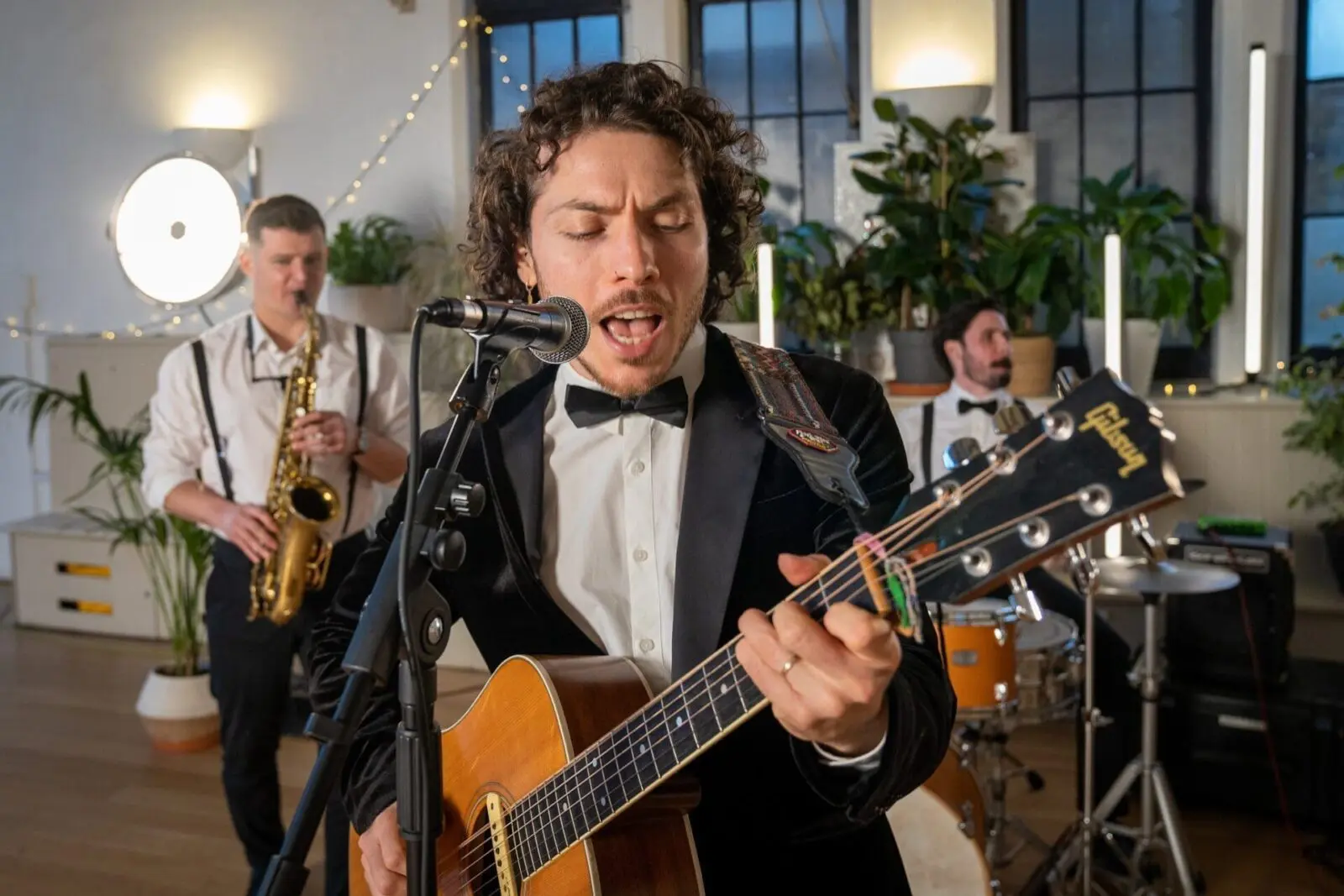 A man in a tuxedo sings and plays acoustic guitar into a microphone, with a saxophonist and drummer performing behind him in a well-lit room with plants.