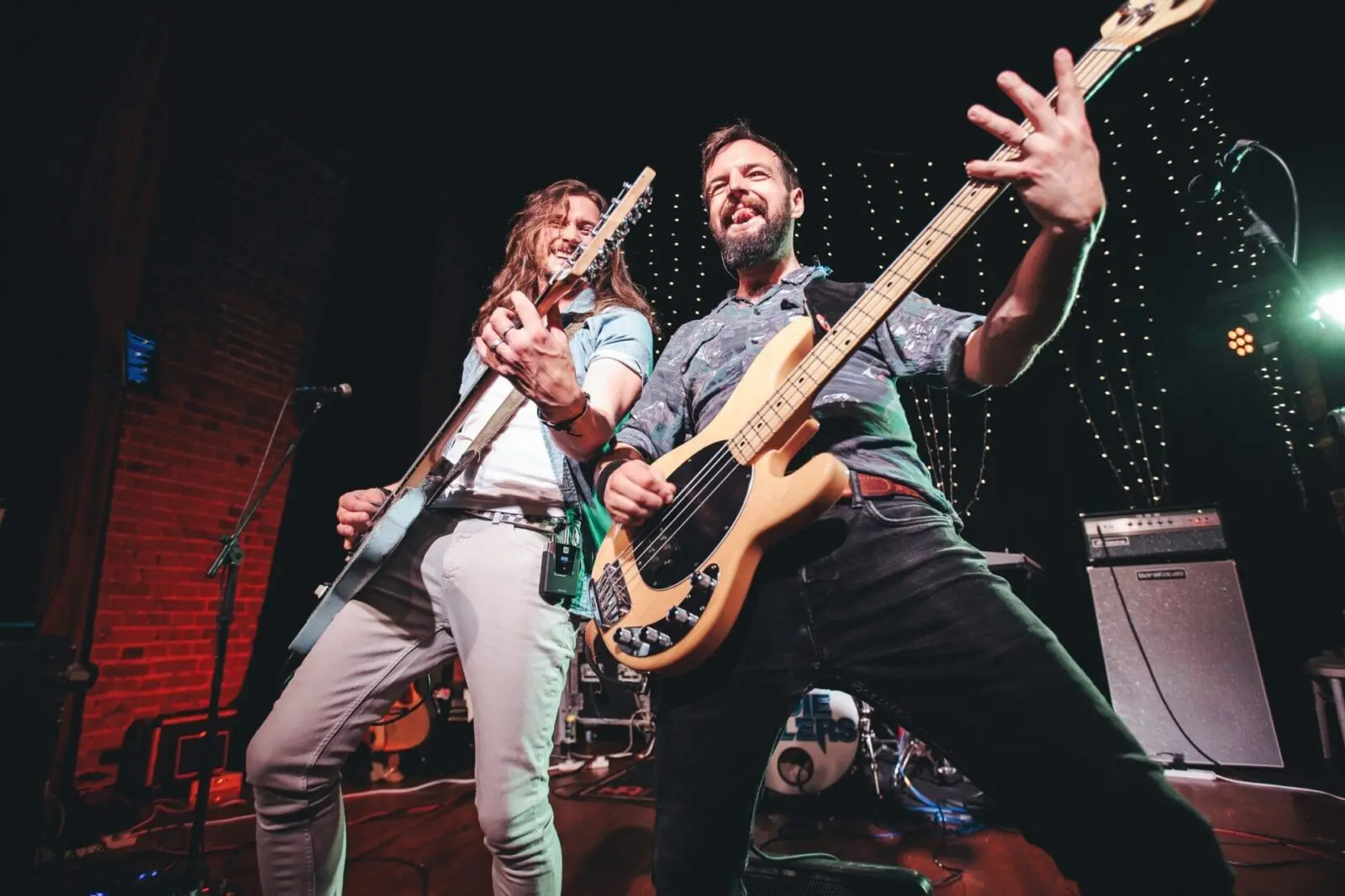 Two musicians playing electric guitars on stage, smiling and posing energetically in front of string lights and musical equipment.