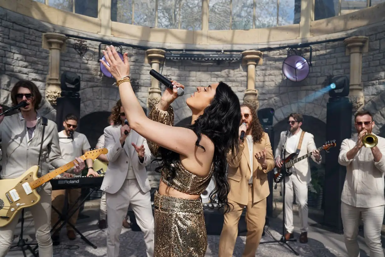 A woman in a gold outfit sings passionately into a microphone while a band in light-colored suits plays various instruments in the background.