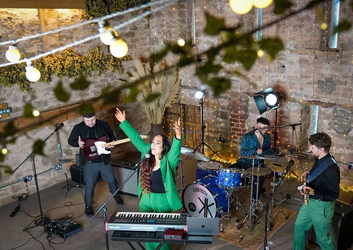 A band performs indoors with a keyboardist singing, a guitarist, a bassist, and a drummer. String lights and rustic decor are visible in the background.