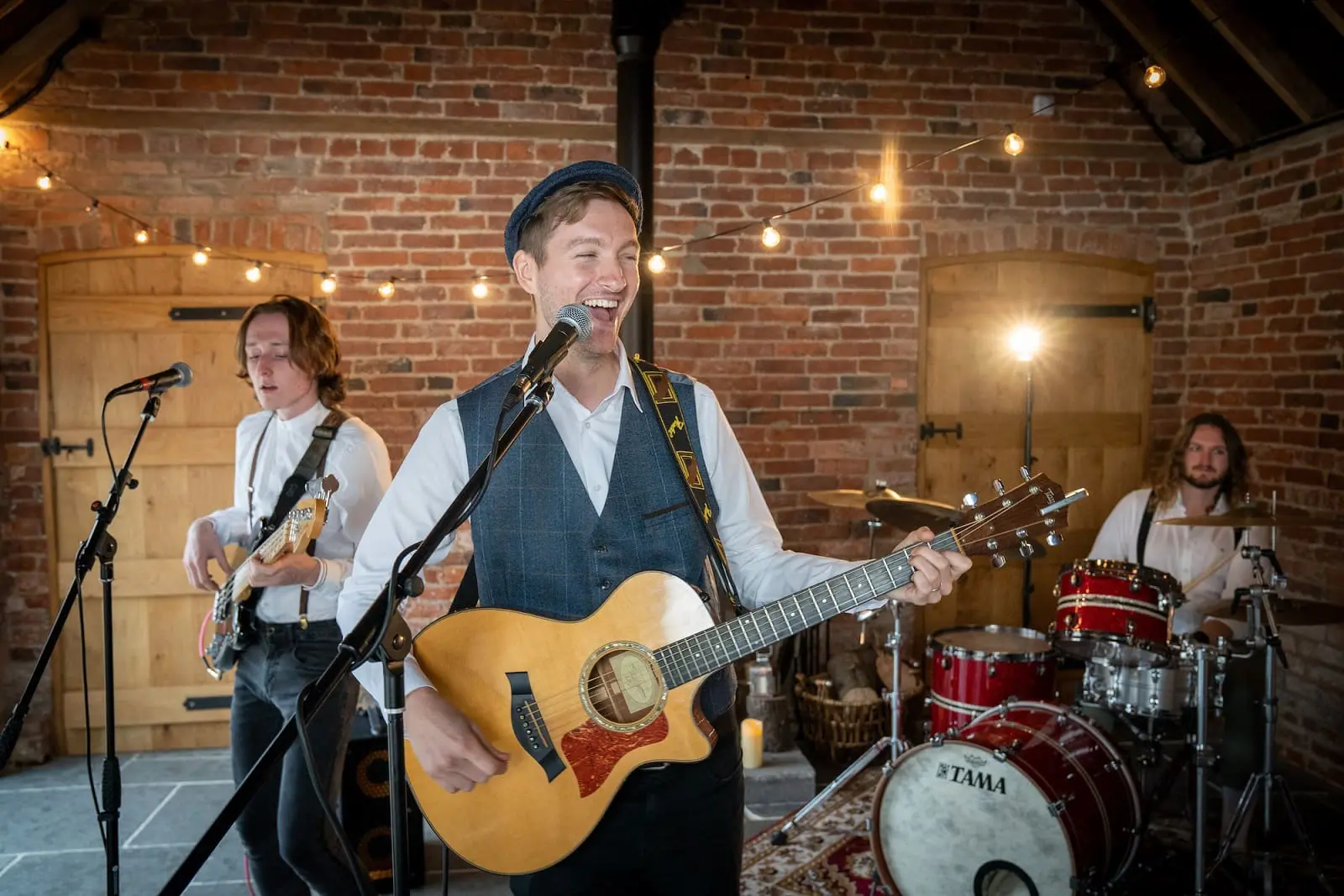 Three musicians perform indoors; the frontman plays acoustic guitar and sings, while another plays bass and one is on drums. String lights and brick walls form the background.