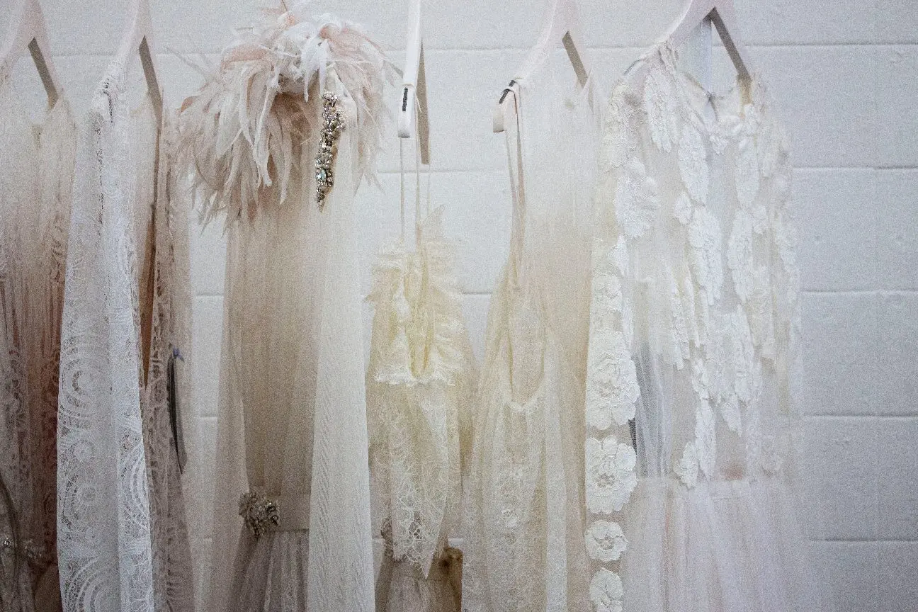 A row of white and cream-colored lacy dresses and outfits hangs on wooden hangers against a white wall.