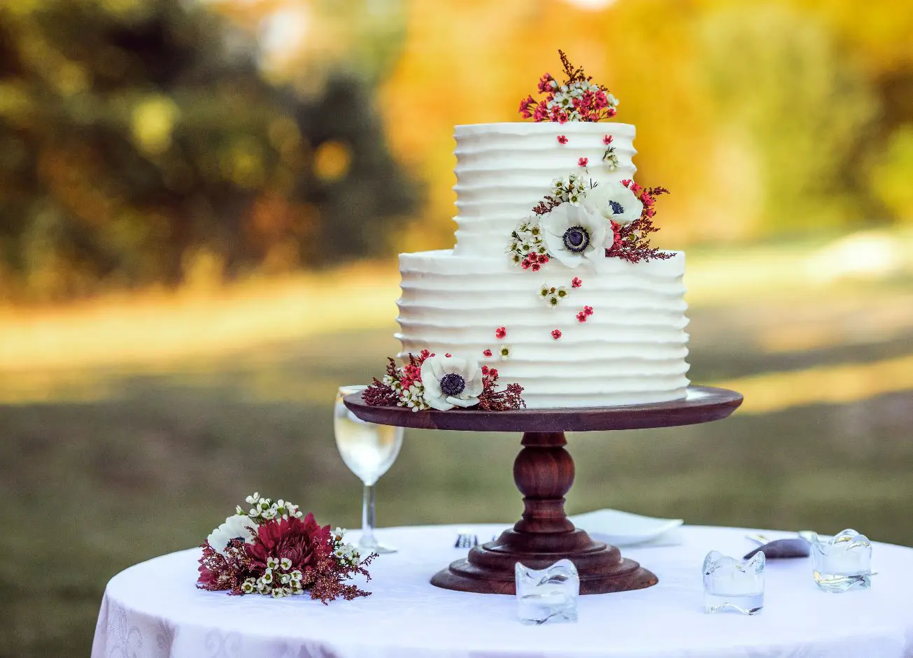 A two-tiered white cake with floral decorations stands on a wooden cake stand on an outdoor table next to a wine glass and a small flower arrangement.