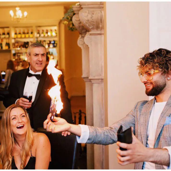 A man in a suit holds a flaming wallet, amusing a seated woman and a standing man at the elegant wedding, adding unexpected flair to an already memorable event.