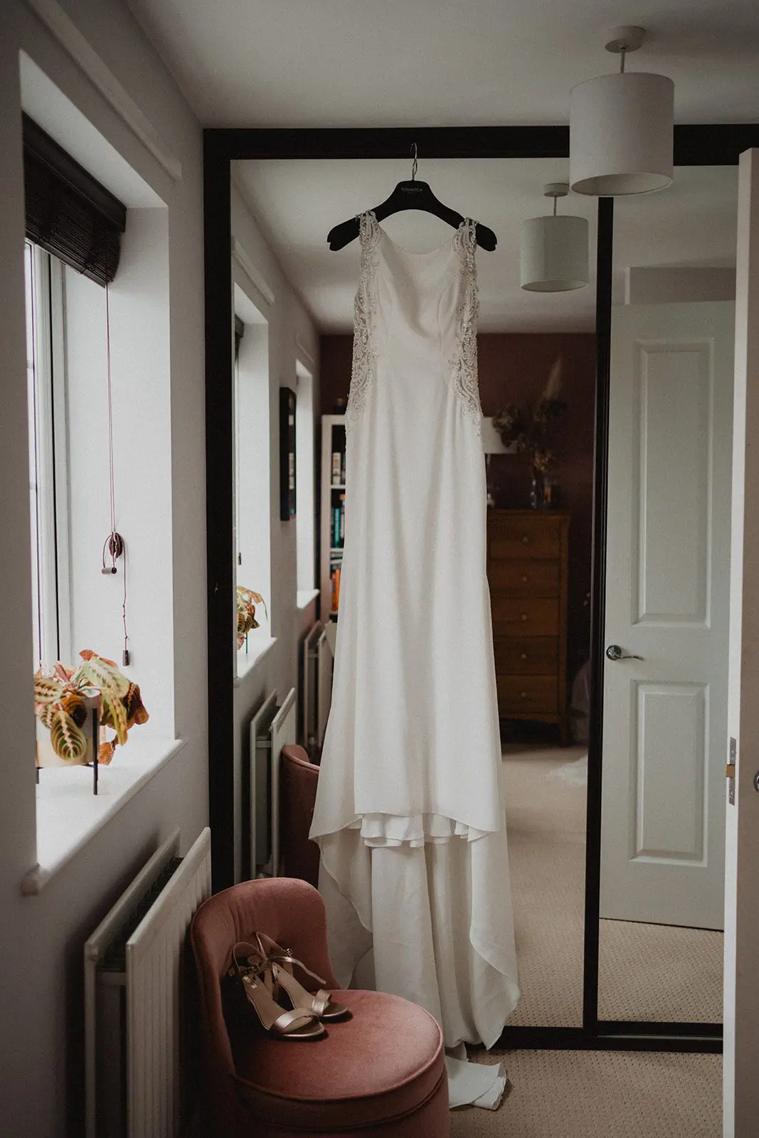 A white dress, perfect for any wedding, hangs on a black hanger in front of a mirror, with beige high heels elegantly placed on a coral chair nearby.