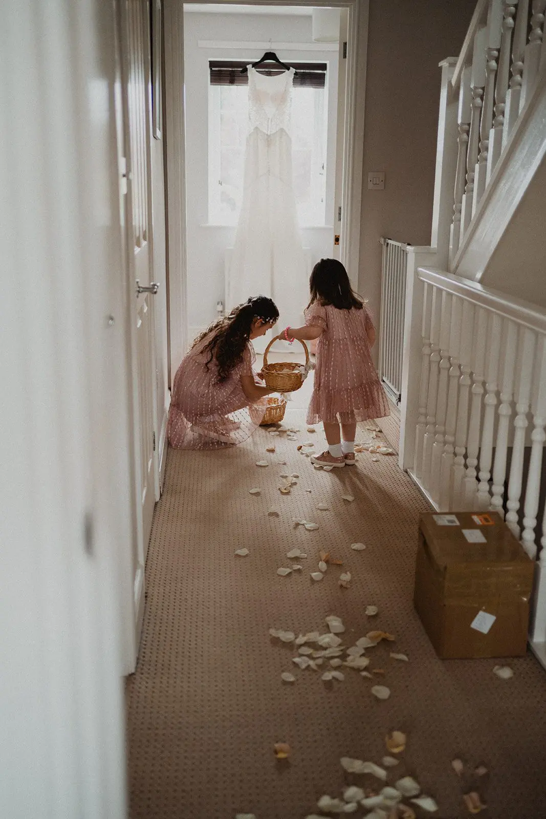 Two children in dresses scatter flower petals down the hallway, a charming prelude to the celebration ahead. In the background, a pristine white gown hints at an enchanting wedding moment soon to unfold.