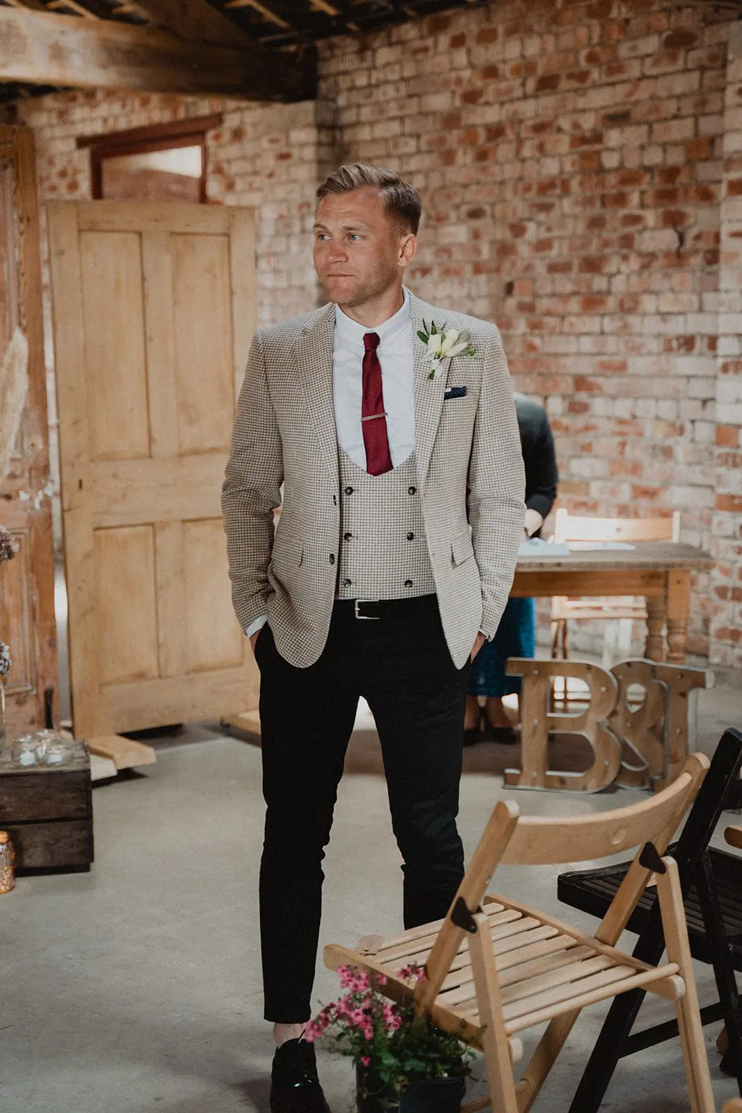 A man in a beige blazer, red tie, and black pants stands indoors, hands in pockets, against a brick wall background. Wood chairs and a large "B" and "J" are in the foreground, hinting at an elegant setup for a wedding service.