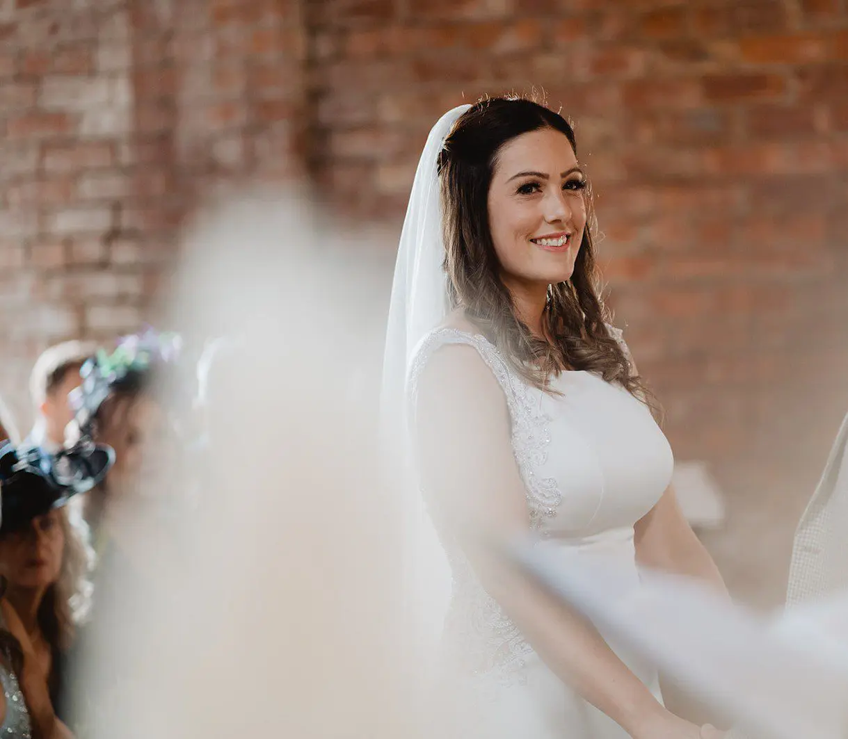 A bride in a white dress and veil stands indoors, smiling, with a blurred brick wall in the background, showcasing the elegance of wedding services.