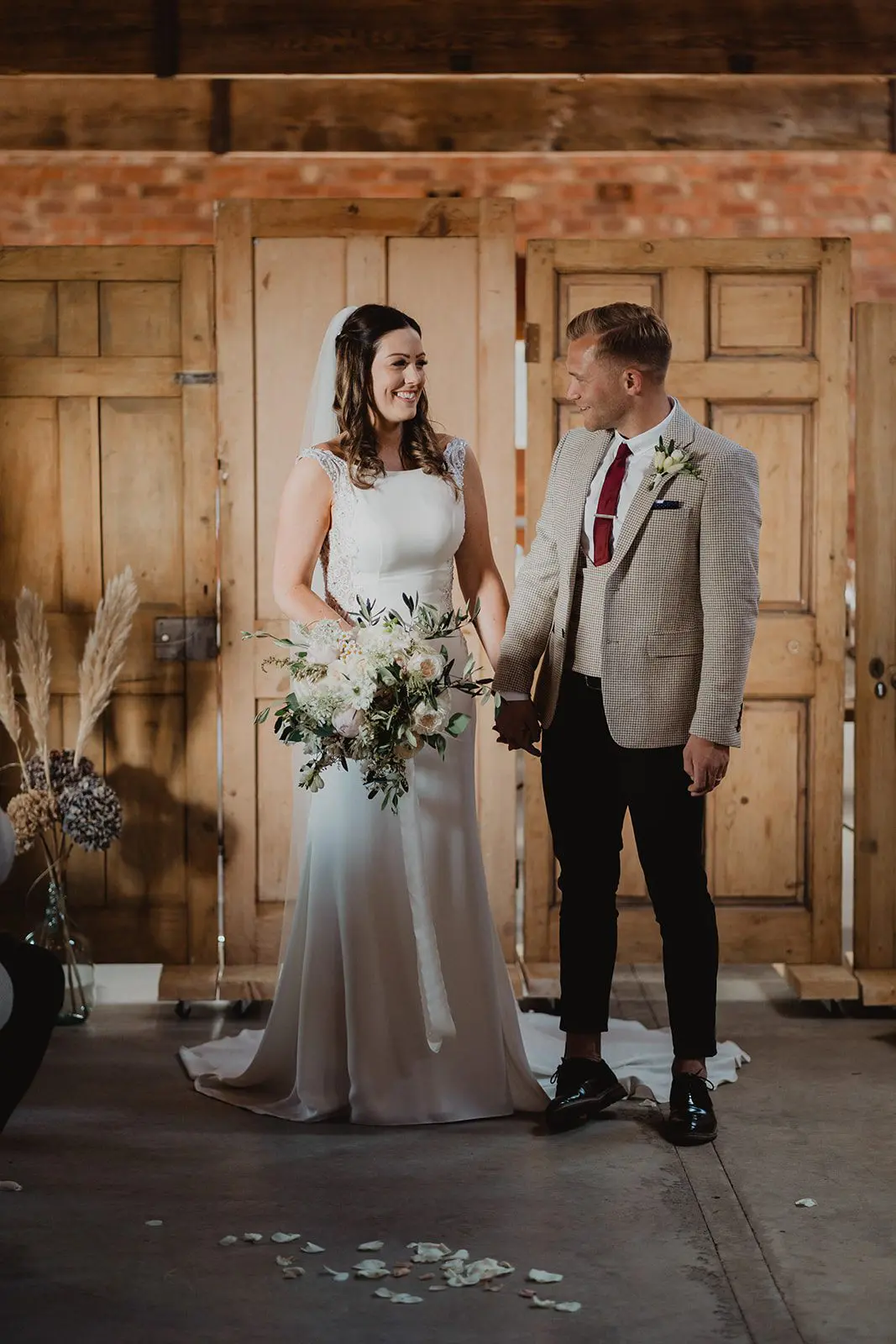A bride and groom stand in front of wooden doors, a testament to flawless wedding planning. The bride holds a bouquet, and both are elegantly dressed in wedding attire.
