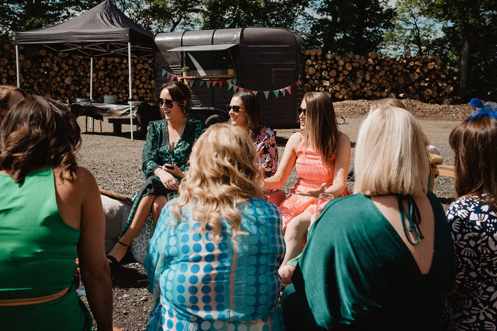 A group of women sit in a circle outdoors, some wearing colorful dresses, with a woodpile and shaded areas in the background. They are deep in discussion about wedding planning, each bringing unique ideas to help shape the perfect day.