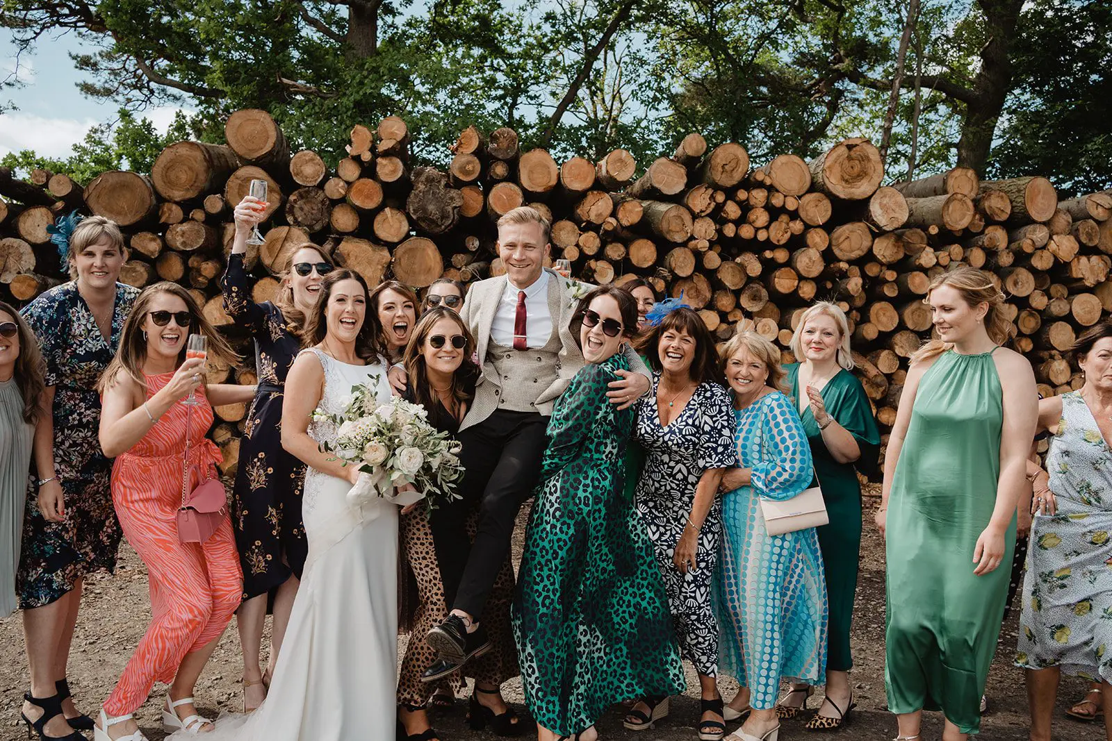 A vibrant group poses in front of stacked logs, with the bride holding a bouquet. The colorful outfits add flair to this joyous scene, reflecting the exceptional work of our wedding planning team.