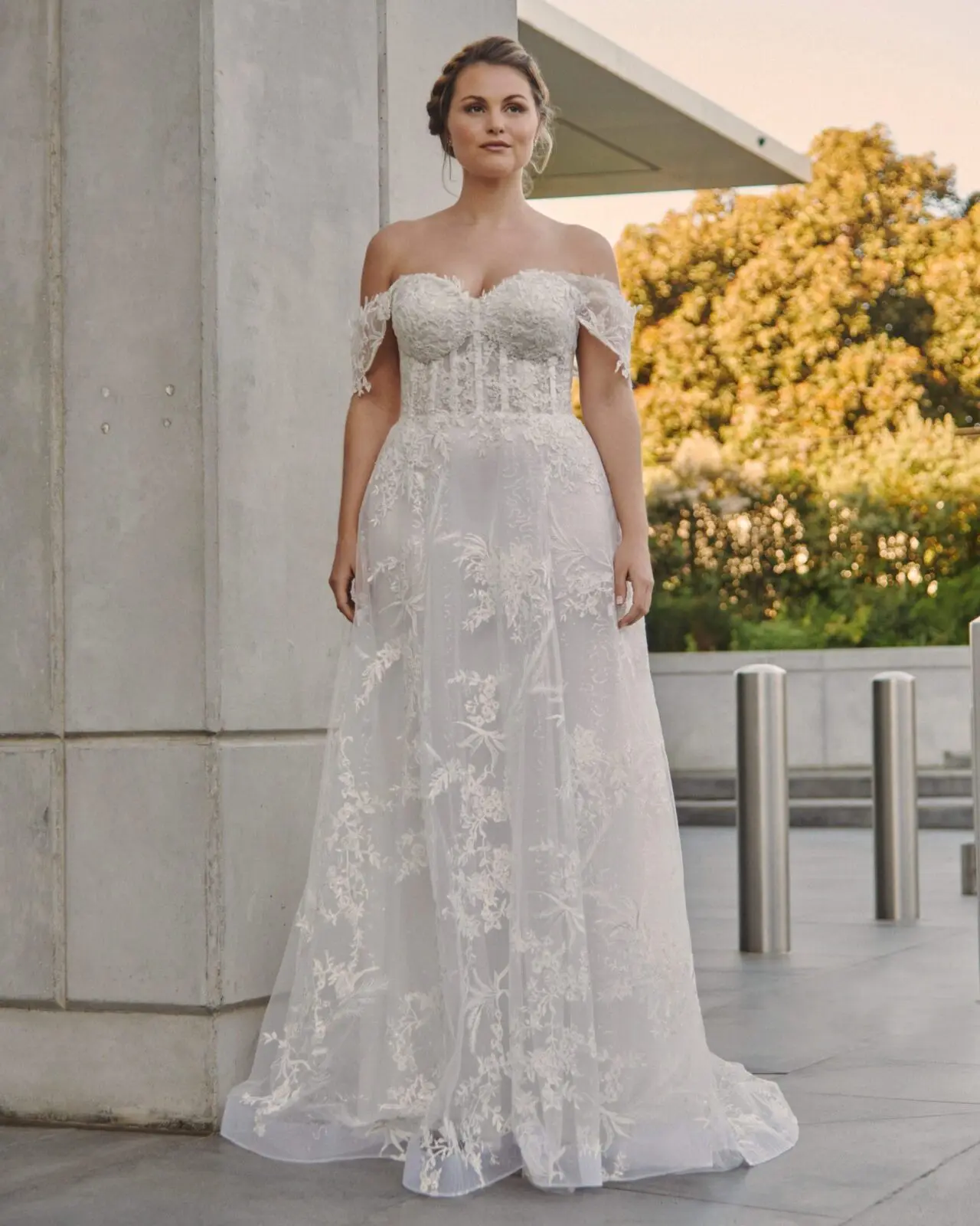 A bride wearing an off-the-shoulder, lace wedding dress stands gracefully outside near a building and lush greenery, the perfect setting that exemplifies exquisite wedding planning in the UK.