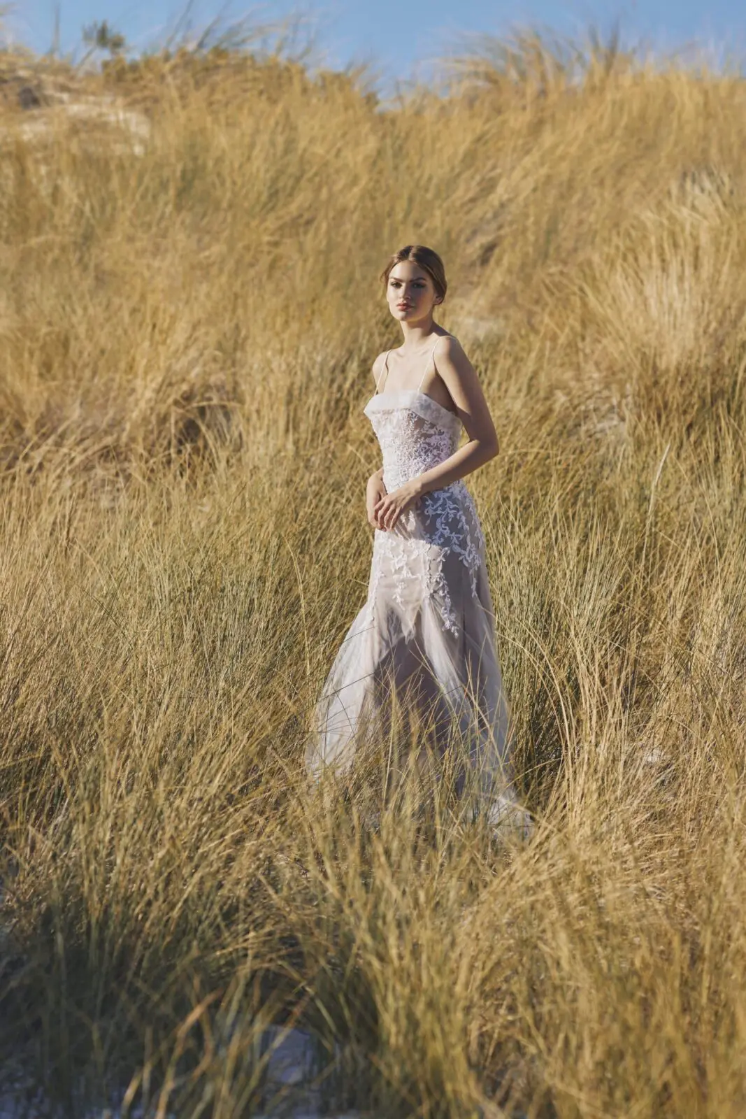 A woman in a white lace dress stands amidst tall golden grass under a clear blue sky, capturing a breathtaking moment that showcases the elegance of our wedding services.