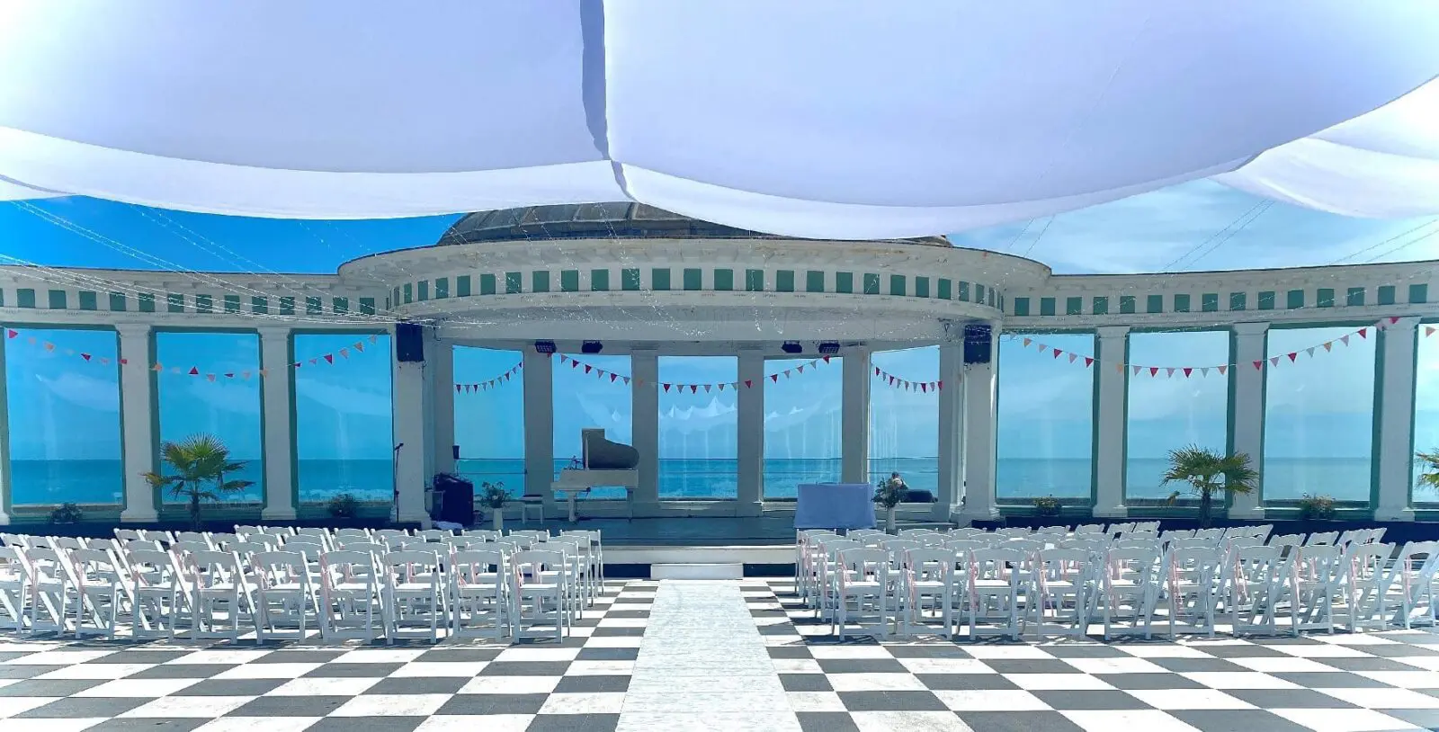 Planning a wedding ceremony in this glass-walled venue is a dream, featuring rows of white chairs on a checkered floor and a grand piano. The ocean view serves as a stunning backdrop, complemented by bunting decorations overhead, perfect for any wedding business looking to showcase elegance.