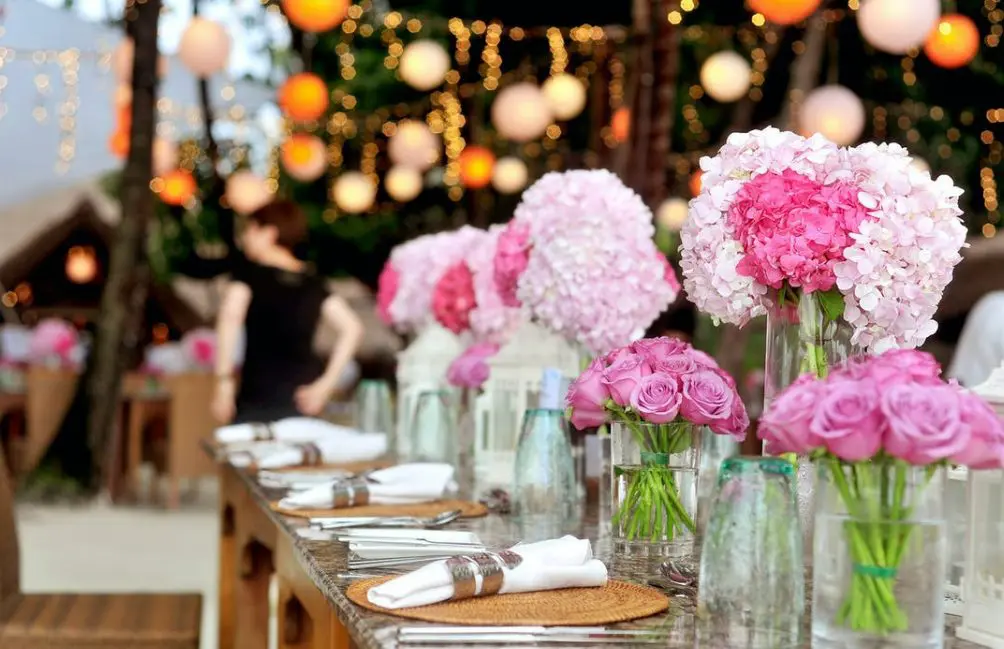 The outdoor dining table is elegantly decorated with pink and purple flower arrangements, ideal for any wedding services. Lanterns and string lights twinkle in the background, creating a magical setting perfect for clients of your wedding business.
