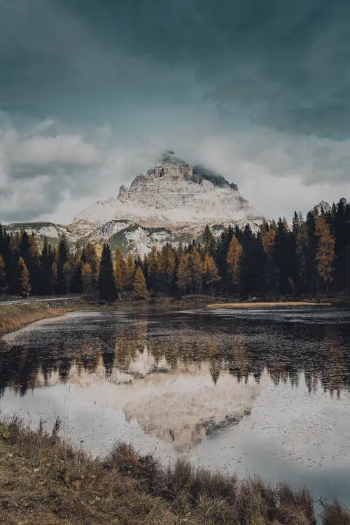 A snow-capped mountain towers over a dense forest, with a calm lake mirroring the breathtaking scenery beneath a cloudy sky. It's an idyllic backdrop for any wedding, where nature's grandeur meets serene tranquility, perfect for couples planning their unforgettable day.
