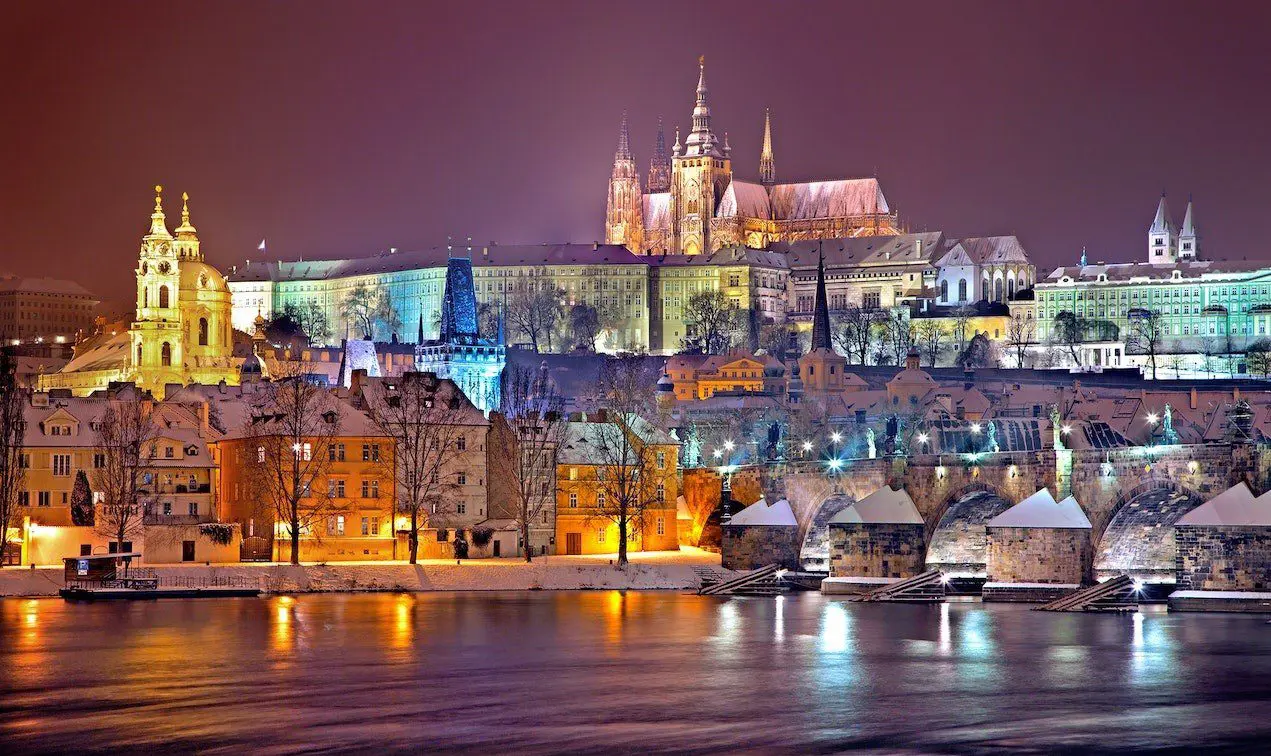 Night view of a brightly lit castle and surrounding historic buildings reflected in a river, with a snowy foreground; an enchanting setting perfect for capturing unforgettable wedding moments.
