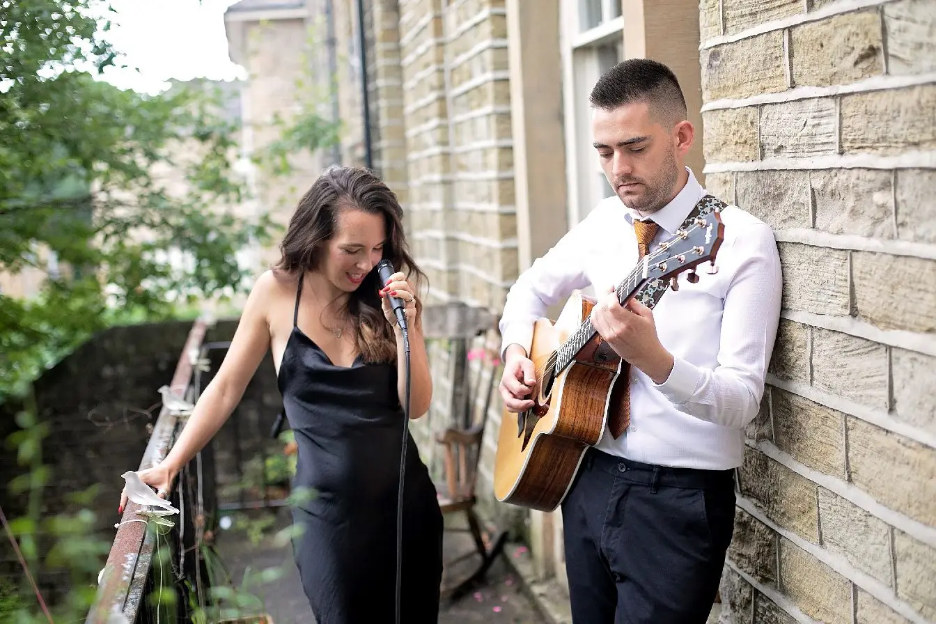 A woman in a black dress sings into a microphone, while a man in a white shirt plays an acoustic guitar on a balcony with brick walls in the background, setting the perfect scene for intimate wedding services.