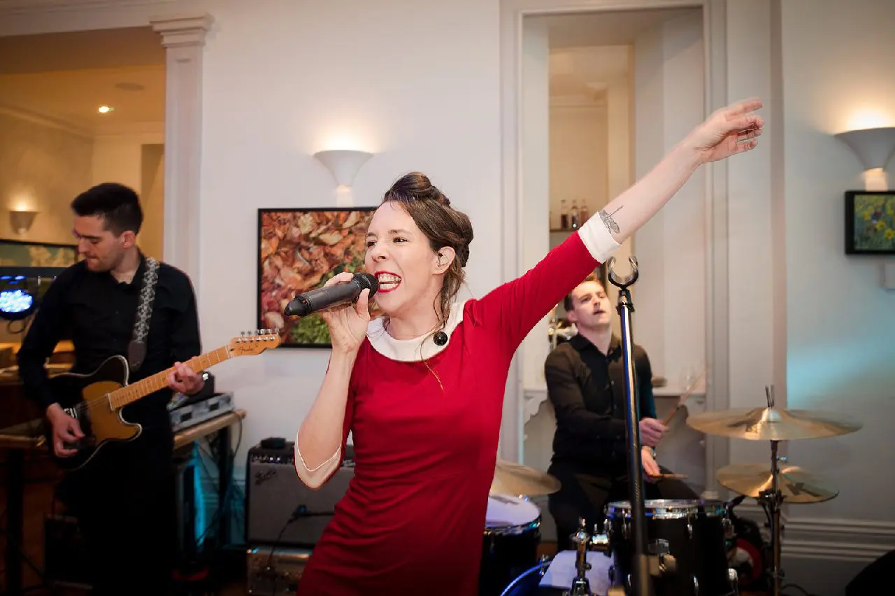 A woman in a red dress sings into the microphone, raising one arm with captivating elegance, as two musicians play guitar and drums in the background. The vibrant performance adds an unforgettable touch to any wedding celebration, seamlessly blending romance and rhythm.