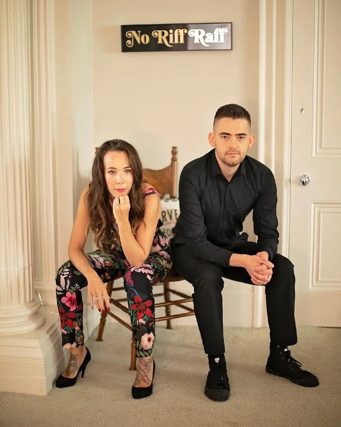 A woman and a man sit on chairs in a room adorned with a sign reading "No Riff Raff" behind them, as they immerse themselves in planning their new wedding business venture.