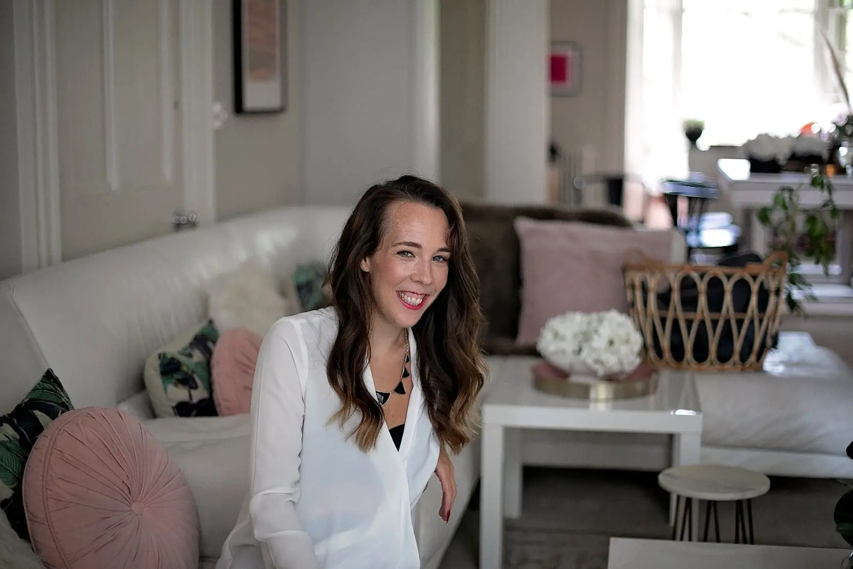 A person smiling and sitting on a white sofa in a stylish, well-decorated living room with pink and black accents, perfectly embodying the elegance often sought in wedding planning.