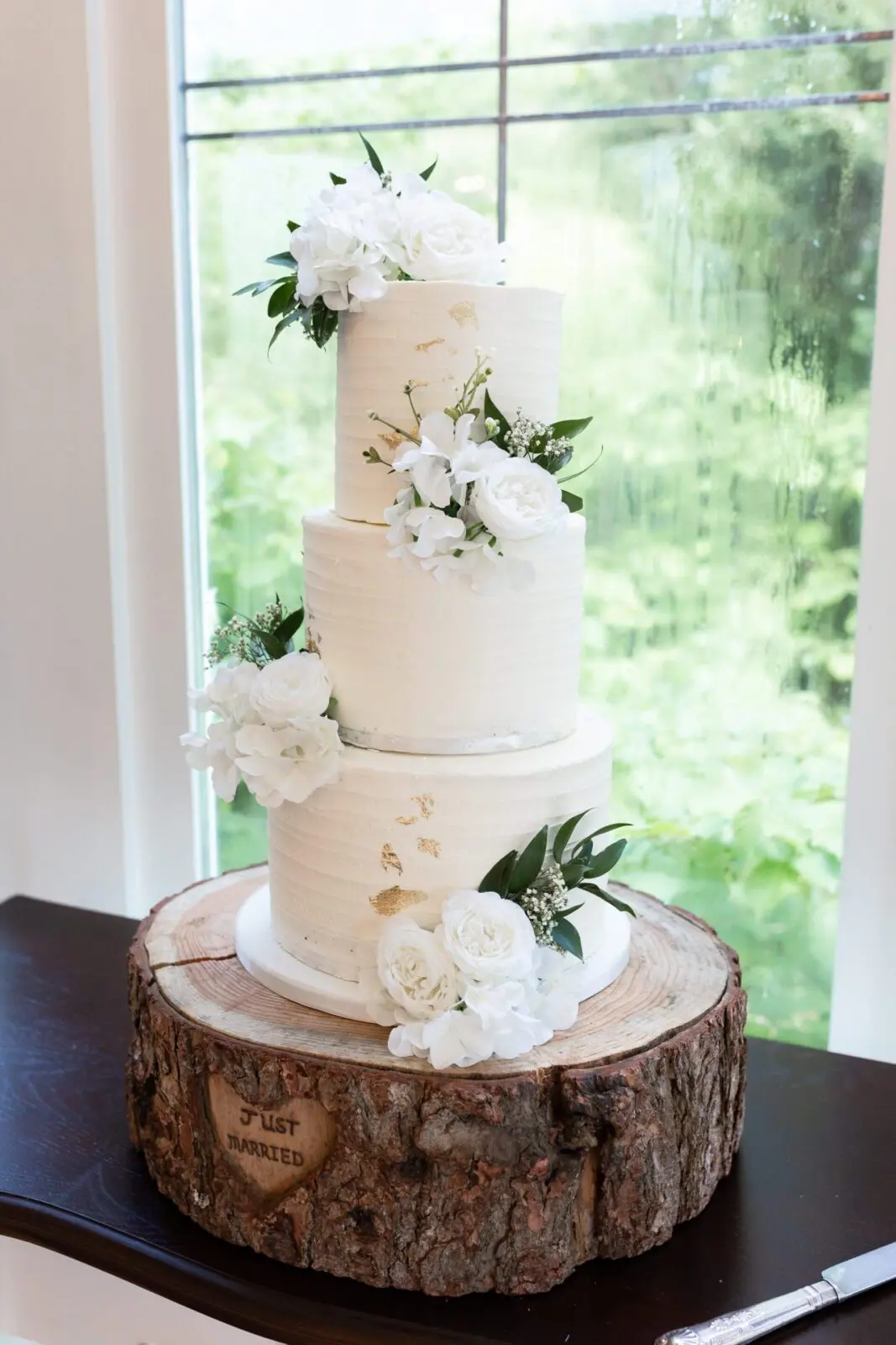 A three-tier white wedding cake with floral decorations, placed on a wooden base inscribed with "JUST MARRIED," captures the essence of elegant wedding planning, set by a rain-speckled window.