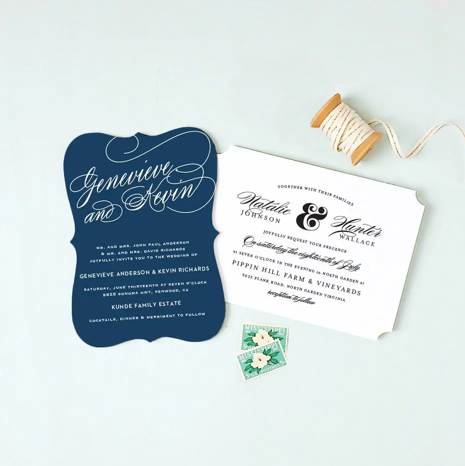 Two wedding invitations in elegant script: one dark blue with white text, the other white with black text. Nearby sits a spool of beige ribbon and two green stamps, perfect for adding a touch of charm to your UK wedding planning essentials.