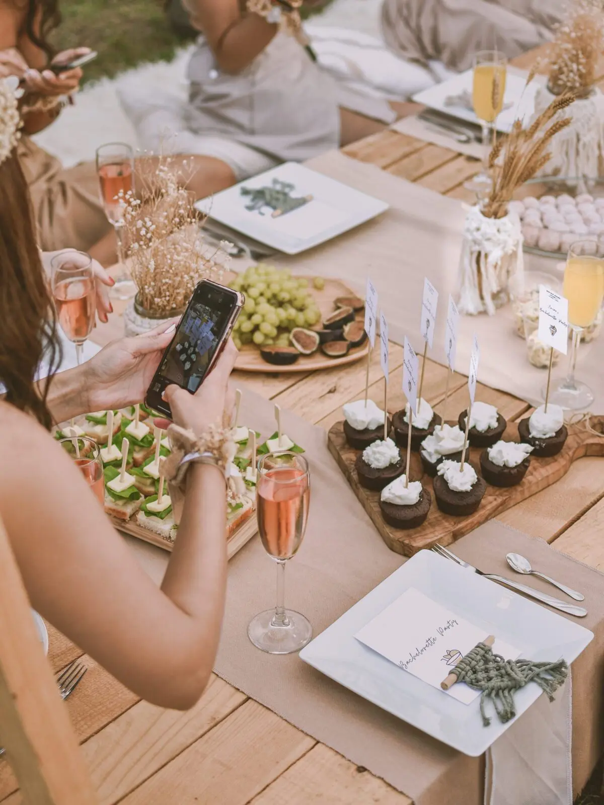 A person takes a photo of a beautifully planned table set with cupcakes, fruit, cocktails, and place cards at an outdoor gathering.