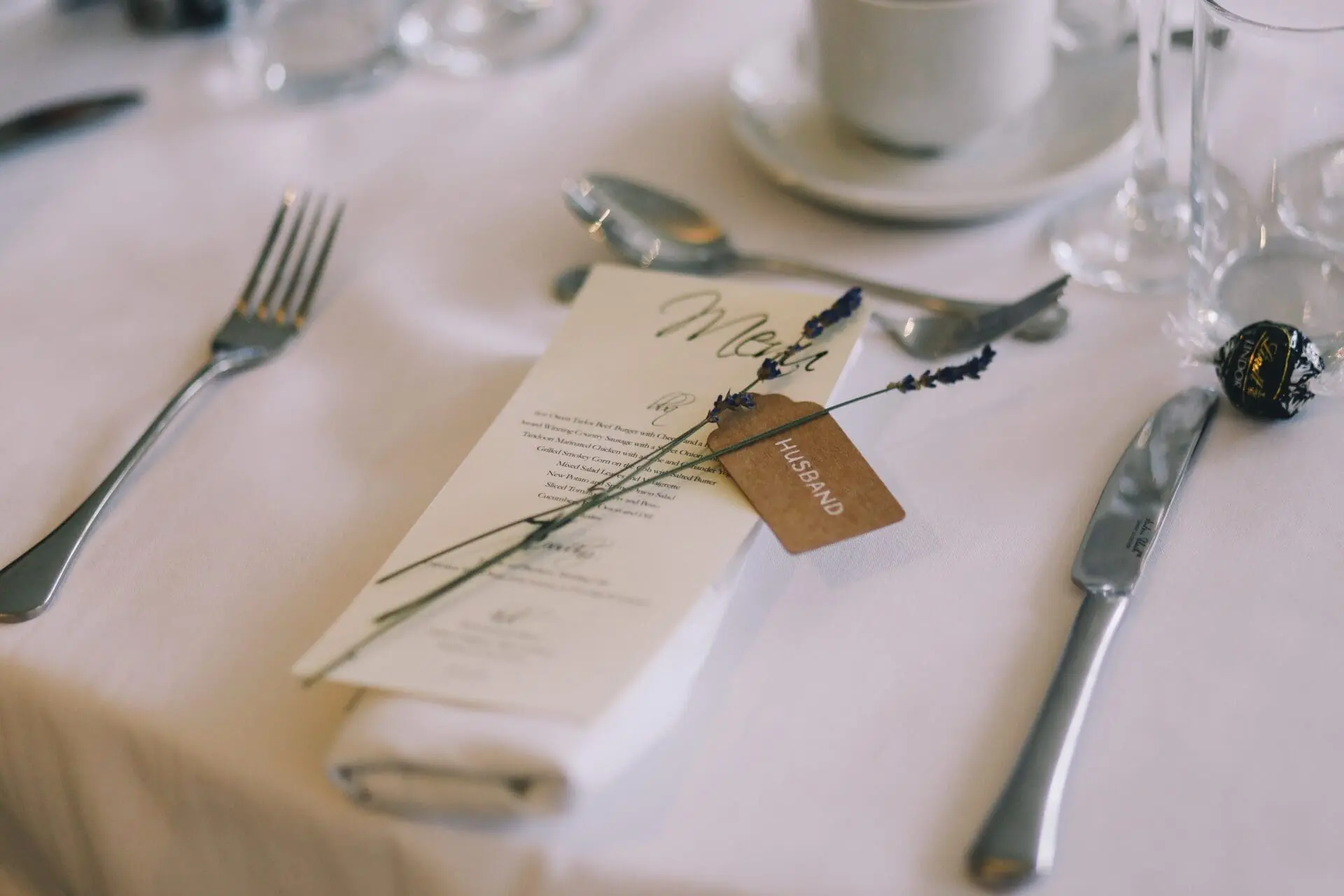 A beautifully planned table setting features a menu, lavender sprig, and a "Husband" tag on a napkin. Silverware, teacup, and glasses are elegantly arranged on a crisp white tablecloth, capturing the essence of thoughtful wedding planning.