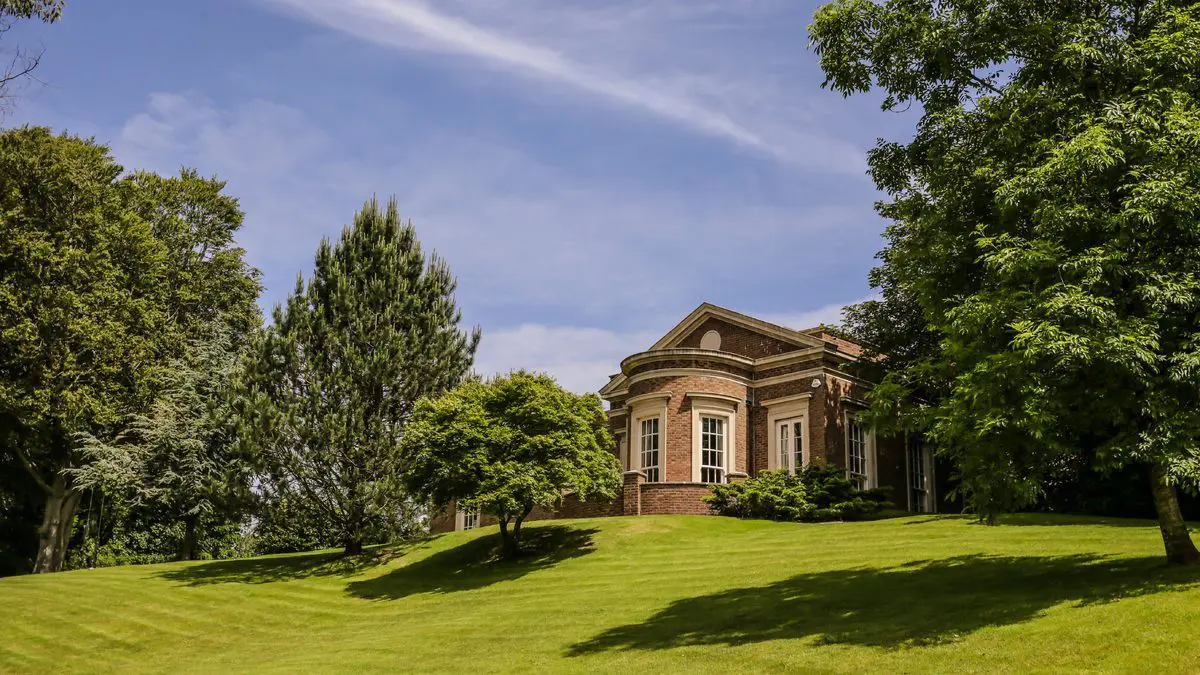 A brick building with large windows, perfect for a wedding business, is surrounded by green grass and trees under a blue sky with wispy clouds.