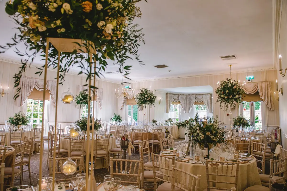 Elegant banquet hall with round tables set for a wedding event. Tables have floral centerpieces, and chairs are arranged around them. The room features large windows and ornate curtains, perfect for wedding planning or any celebration within the flourishing wedding business.