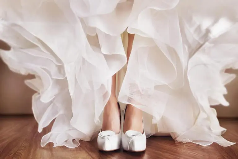 A close-up captures a person in elegant white high-heeled shoes and a flowing ruffled dress, perfect for wedding planning in the UK, standing gracefully on a wooden floor.